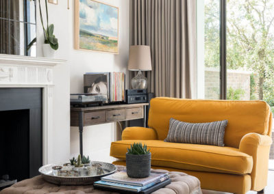 Penman Interiors Dulwich Living Room Ochre Loveseat Buttoned Ottoman Fireplace Black Gold Wall Light Linen Curtains